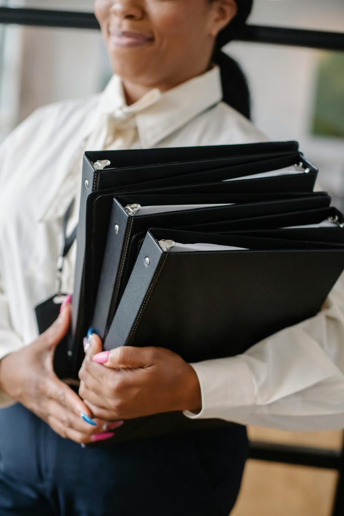 Professional woman in formal attire carrying multiple black folders indoors.