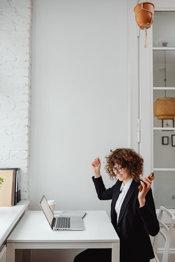 Businesswoman with curly hair celebrates success on phone call in modern office with laptop.